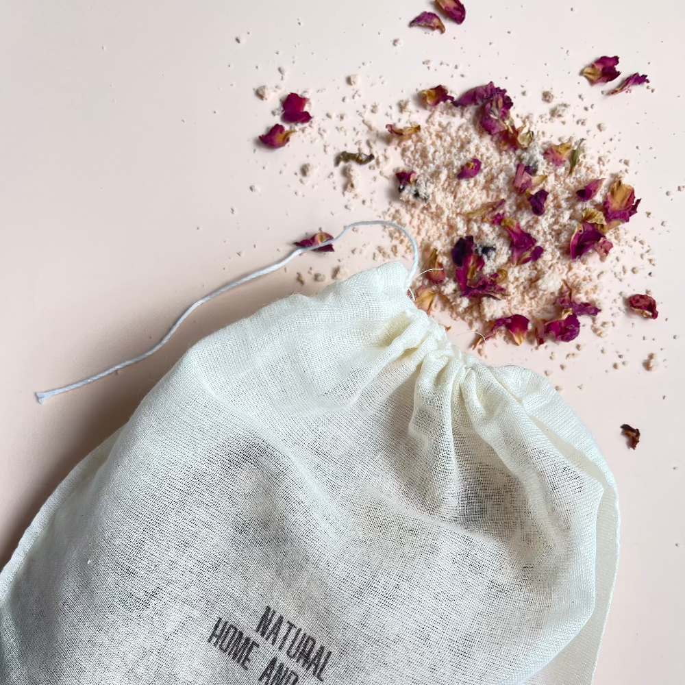 Bath bag in action with some salts and rose petals coming out of the bag on a pink background.