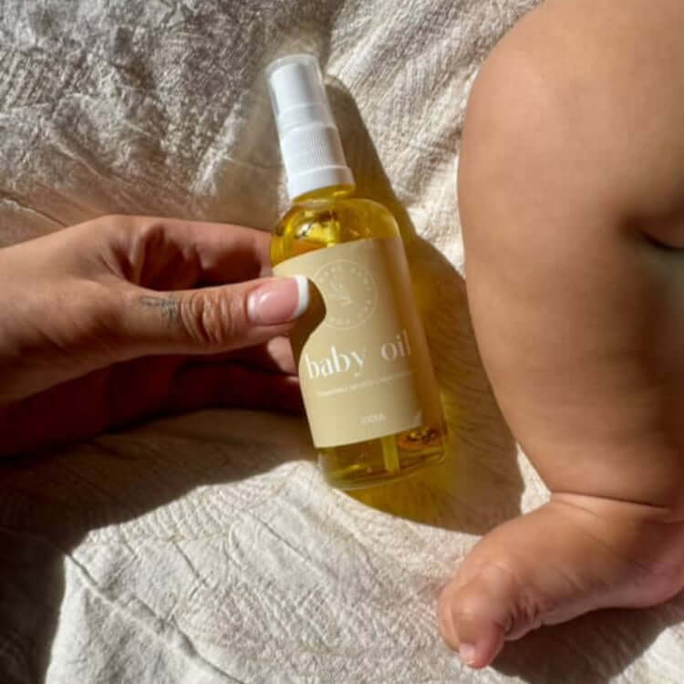 Organic baby oil centred in the middle with a hand coming into the left and holding the bottle, from a birds eye view. A baby leg is next to the bottle, on the right hand side.