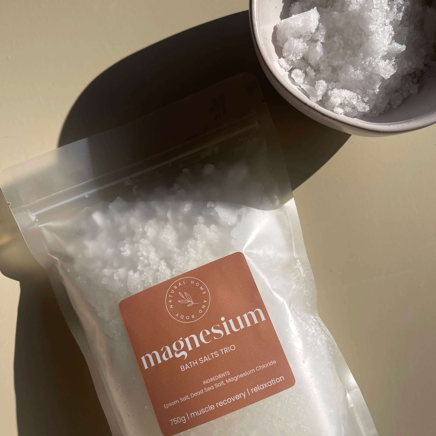 Magnesium bath salts bag displayed on a beige background . At the top right of the image, is a bowl filled with the bath salts. The sun is shining in displaying shadows.