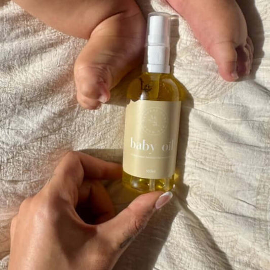 Organic Baby Oil on a neutral bed sheet. Mums hand holding the bottom of the bottle and baby feet entering the top of the image.